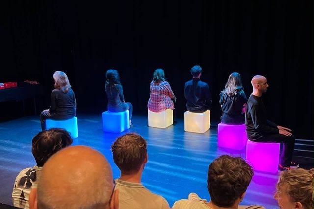 The cast seated on glowing blue, amber and magenta cubes, seen from the audience